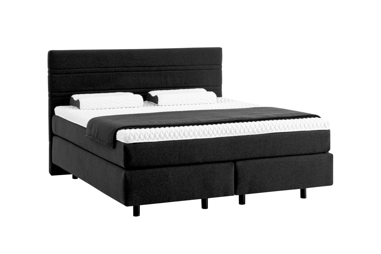Boxspringbett in dunklem Stoffbezug mit zwei Kissen und einer Decke, seitliche Perspektive