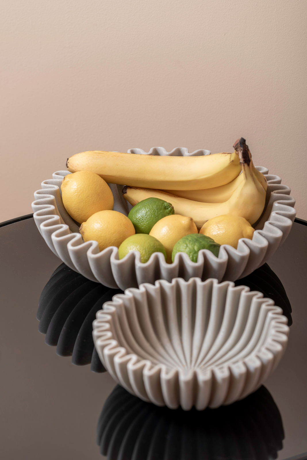 Zwei graue Schalen mit gewelltem Rand, gefüllt mit Bananen, Zitronen und Limetten, auf einem schwarzen Tisch aus der Vogelperspektive.