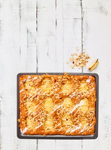 Vogelperspektive auf ein Backblech mit einem Apfelkuchen, dekoriert mit Mandelscheiben und Zuckerguss, auf einem weißen Holztisch.
