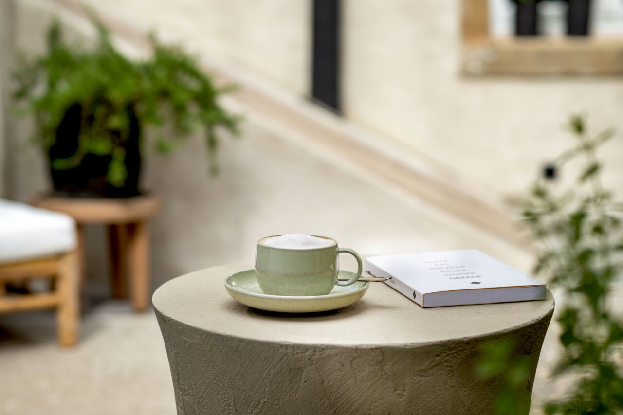 Grüne Kaffeetasse mit Untertasse auf einem Tisch, daneben ein Buch, im Hintergrund Pflanzen. Perspektive: seitlich von oben.