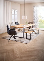 Modernes Esszimmer mit rechteckigem Esstisch aus hellem Massivholz und schwarzem Metallgestell, passender Holzbank und gepolsterten Stühlen auf Teppich, aufgenommen in leichter Schrägansicht von vorn.