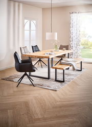 Modernes Esszimmer mit rechteckigem Esstisch aus hellem Massivholz und schwarzem Metallgestell, passender Holzbank und gepolsterten Stühlen auf Teppich, aufgenommen in leichter Schrägansicht von vorn.