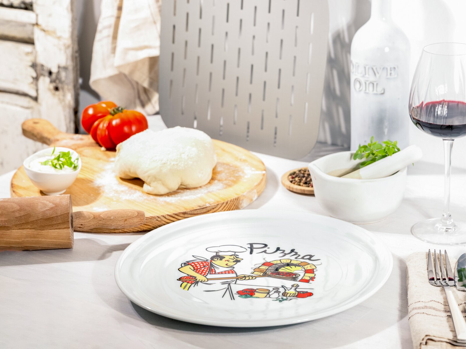 Weißer Pizzateller mit Pizza-Motiv und Schriftzug „Pizza“ auf einem gedeckten Tisch, im Vordergrund aus leicht schräger Draufsicht; im Hintergrund Pizzateig auf Holzbrett, Tomaten, Mörser, Olivenölflasche und Rotweinglas.