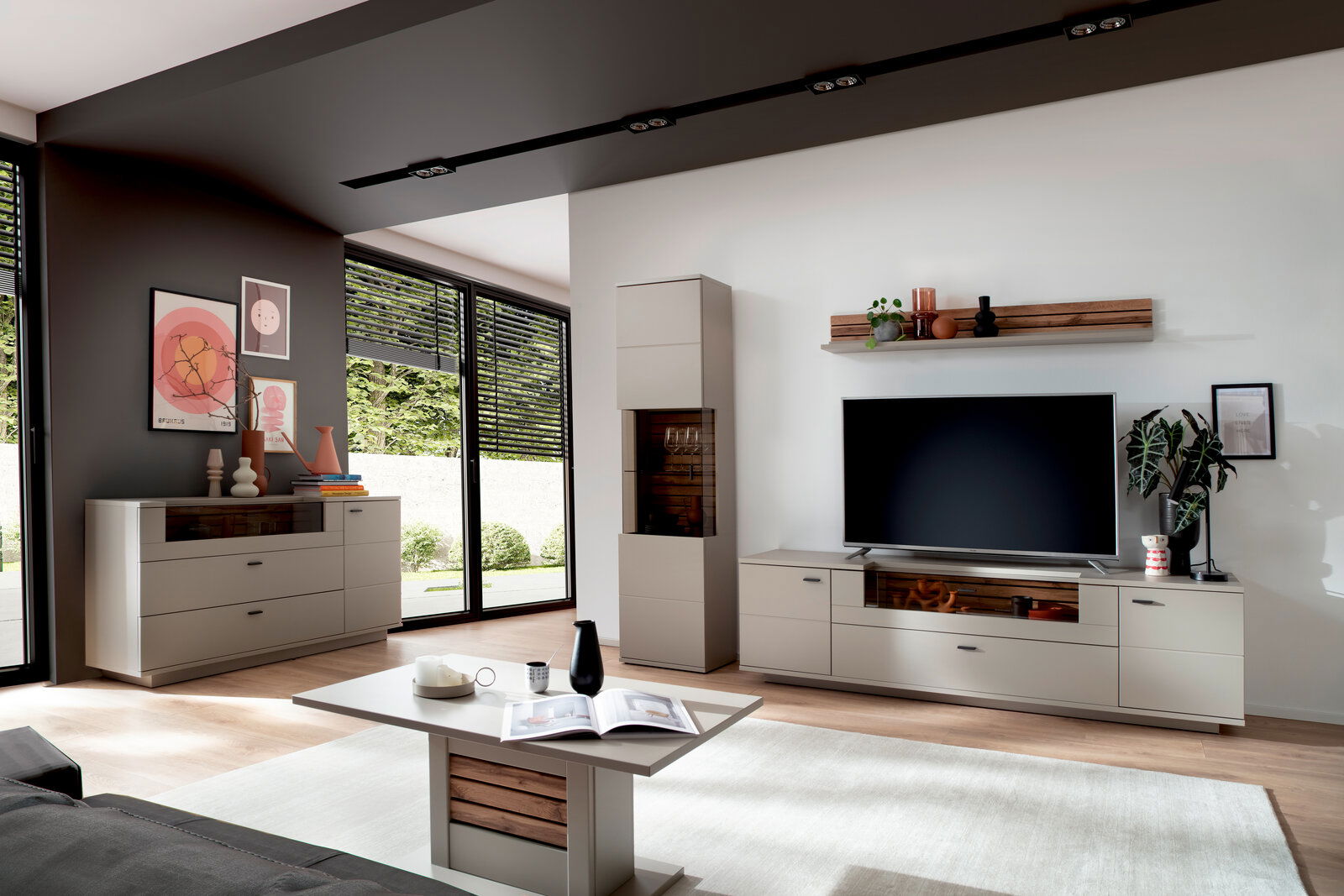 Modernes Wohnzimmer mit hellgrauer Wohnwand und Vitrine mit Holz-Akzenten, Lowboard mit großem Fernseher, Sideboard und Couchtisch; Perspektive: Weitwinkelaufnahme aus leicht erhöhter, frontaler Ecke.