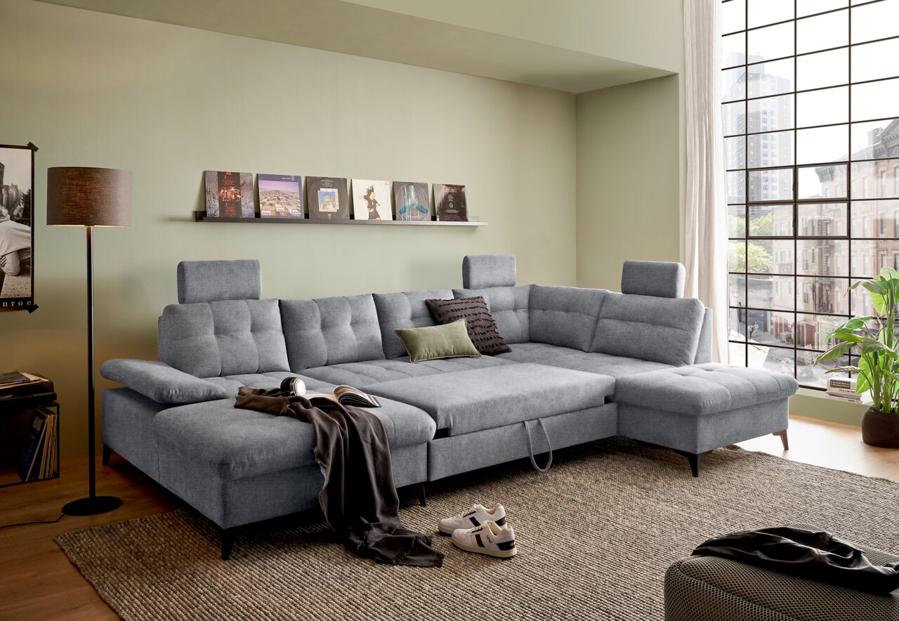 Graues Ecksofa in einem modernen Wohnzimmer, mit Blick von vorne. Das Sofa hat verstellbare Kopfstützen und ist mit Kissen dekoriert. Im Hintergrund sind ein großes Fenster und ein Regal mit Schallplatten zu sehen.