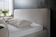 Seitliche Ansicht eines Polsterbettgestells mit hellem, geripptem Kopfteil in einem gemütlichen Schlafzimmer mit Fenster im Hintergrund.