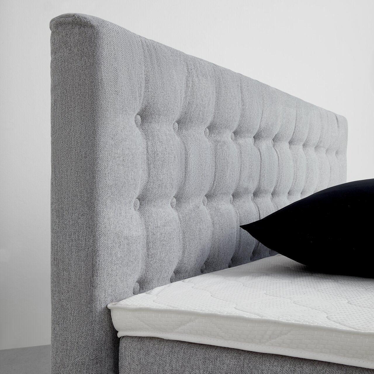 Seitliche Ansicht eines grauen Boxspringbetts mit gestepptem Kopfteil und schwarzem Kissen.