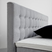 Seitliche Ansicht eines grauen Boxspringbetts mit gestepptem Kopfteil und schwarzem Kissen.