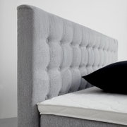Seitliche Ansicht eines grauen Boxspringbetts mit gestepptem Kopfteil und schwarzem Kissen.
