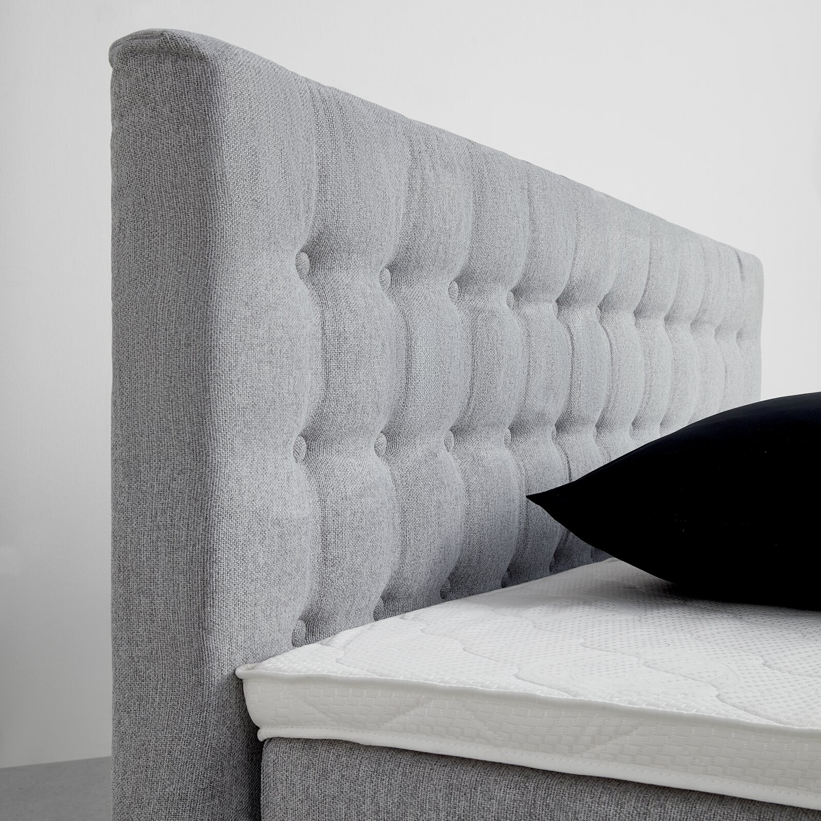 Seitliche Ansicht eines grauen Boxspringbetts mit gestepptem Kopfteil und schwarzem Kissen.
