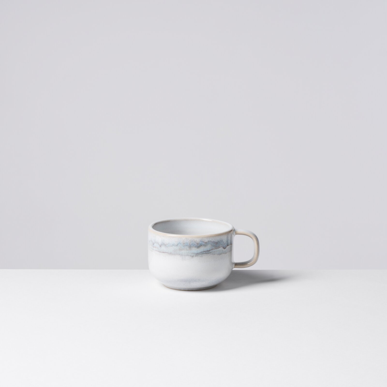 Weiße Keramik-Kaffeetasse mit hellblau-grauem, marmoriertem Streifen und Henkel rechts, freigestellt vor hellem Hintergrund, frontal in leichter Seitenansicht