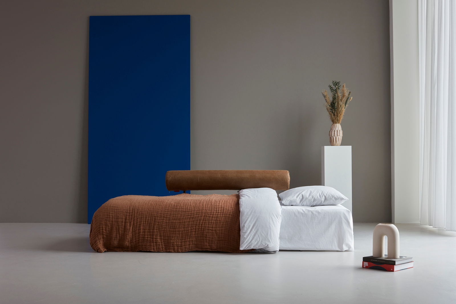 Seitliche Ansicht eines Funktionssofas, das als Bett genutzt wird, mit brauner Decke und weißen Kissen, im Hintergrund eine blaue Wand und eine Vase auf einem Sockel.