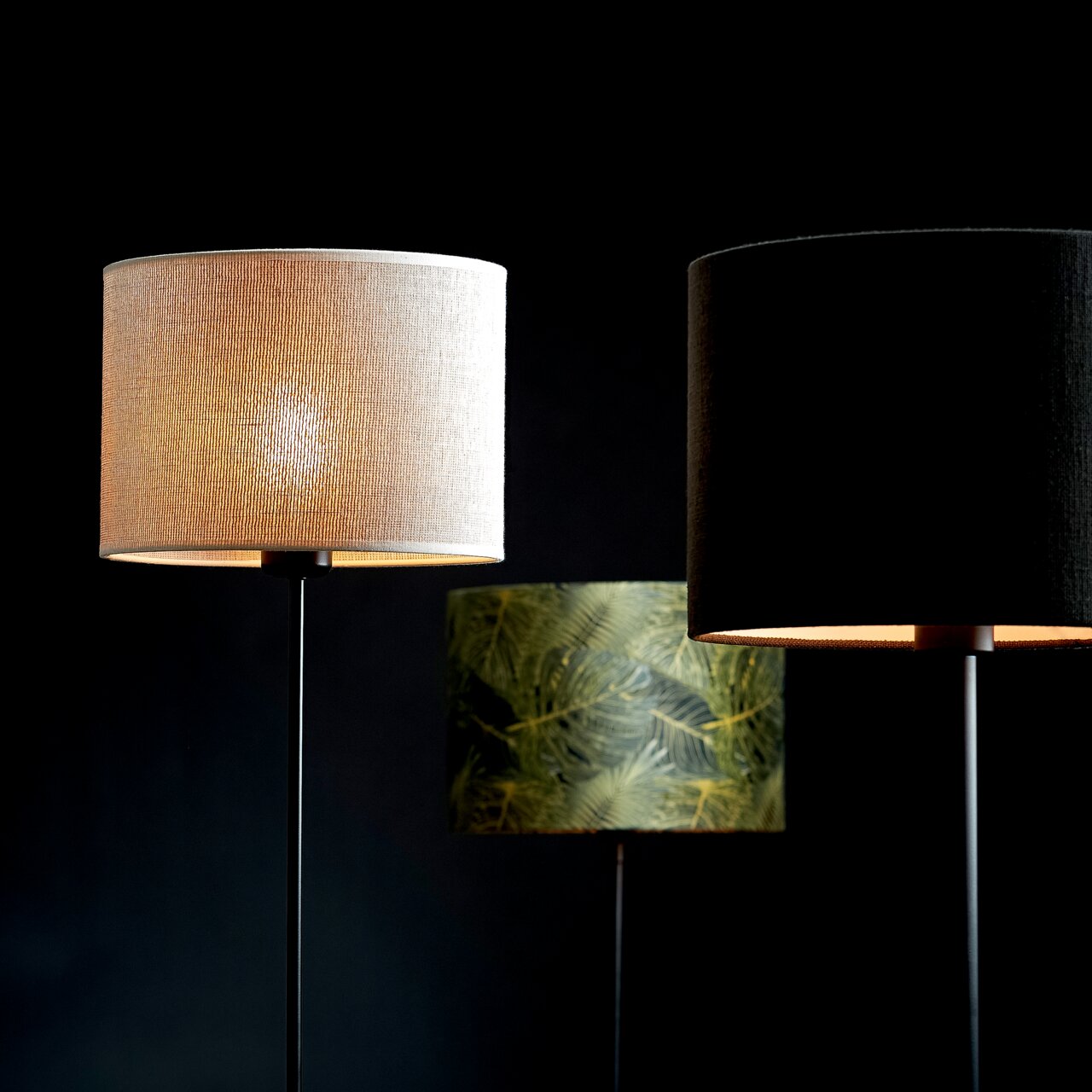 Drei moderne Lampenschirme in verschiedenen Designs, frontal betrachtet: ein heller Stoffschirm, ein schwarzer Stoffschirm und ein grüner Schirm mit Blattmuster.
