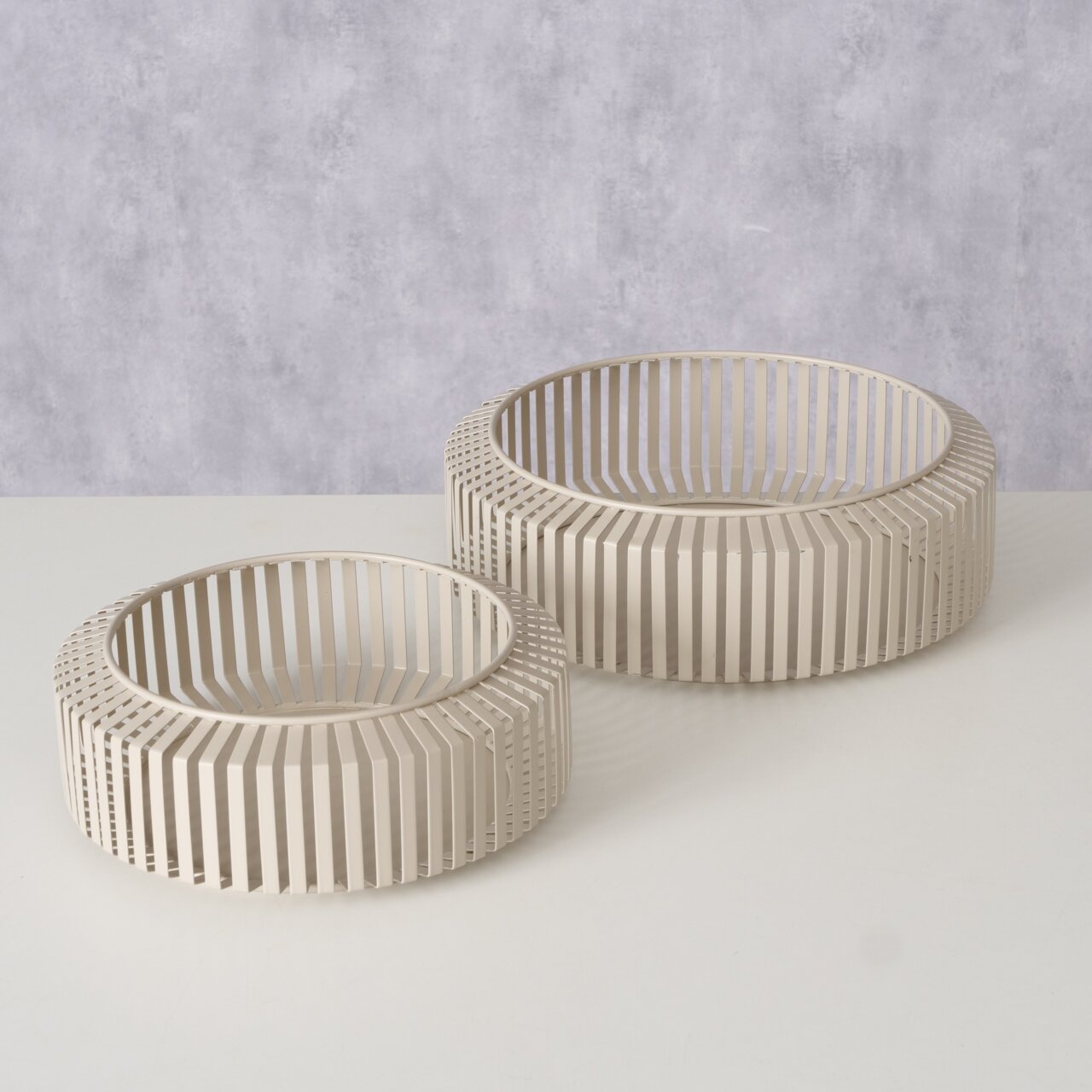 Zwei moderne, runde Schalen aus beigem Metall mit einem offenen, lamellenartigen Design, auf einer weißen Oberfläche aus der Vogelperspektive fotografiert.