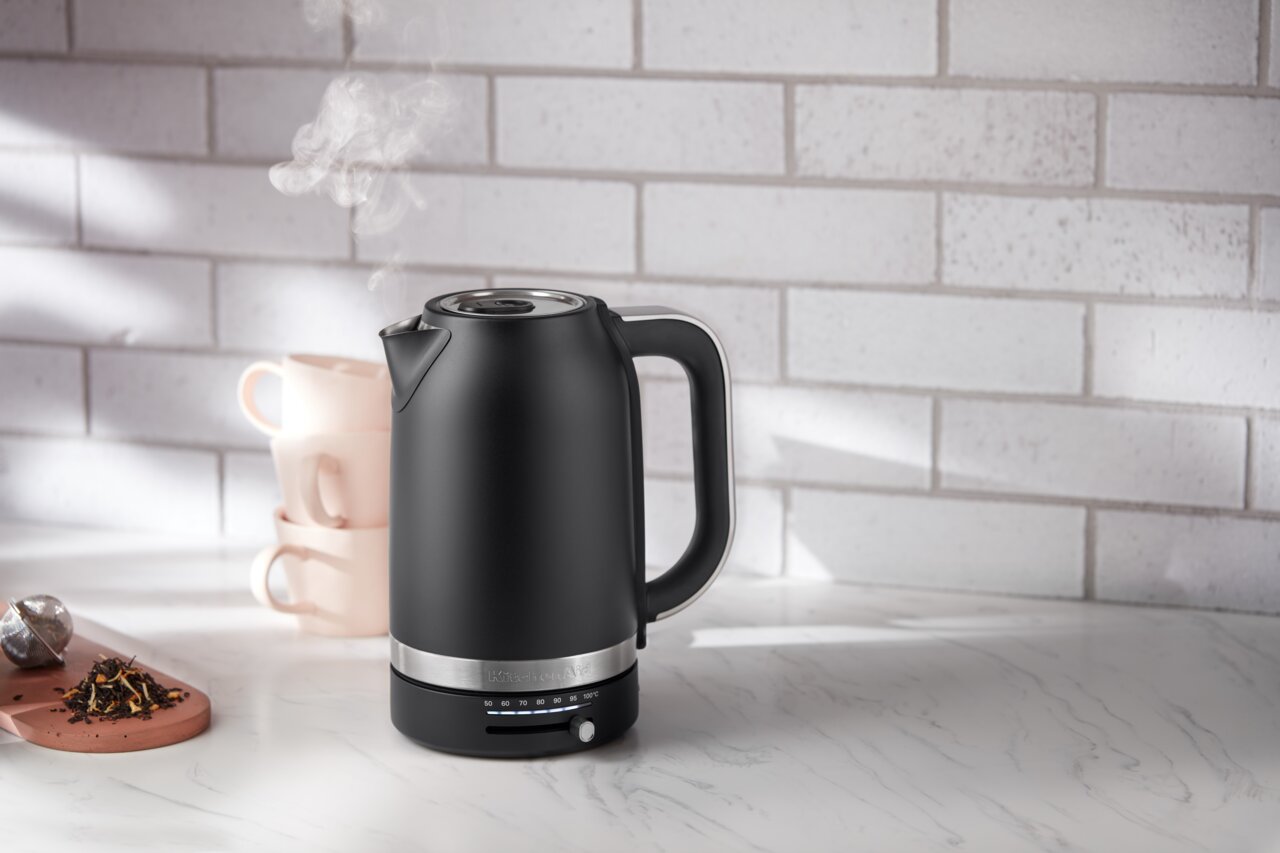 KitchenAid Wasserkocher Schwarzer Wasserkocher mit 1,7 Liter Fassungsvermögen, seitliche Perspektive, auf einer Küchenarbeitsplatte mit Tassen und Tee im Hintergrund.