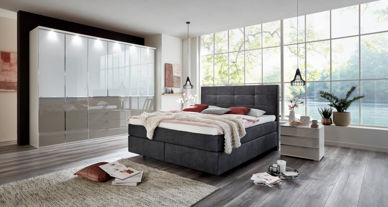 Modernes Boxspringbett in einem stilvollen Schlafzimmer mit großem Fenster im Hintergrund, seitliche Perspektive.