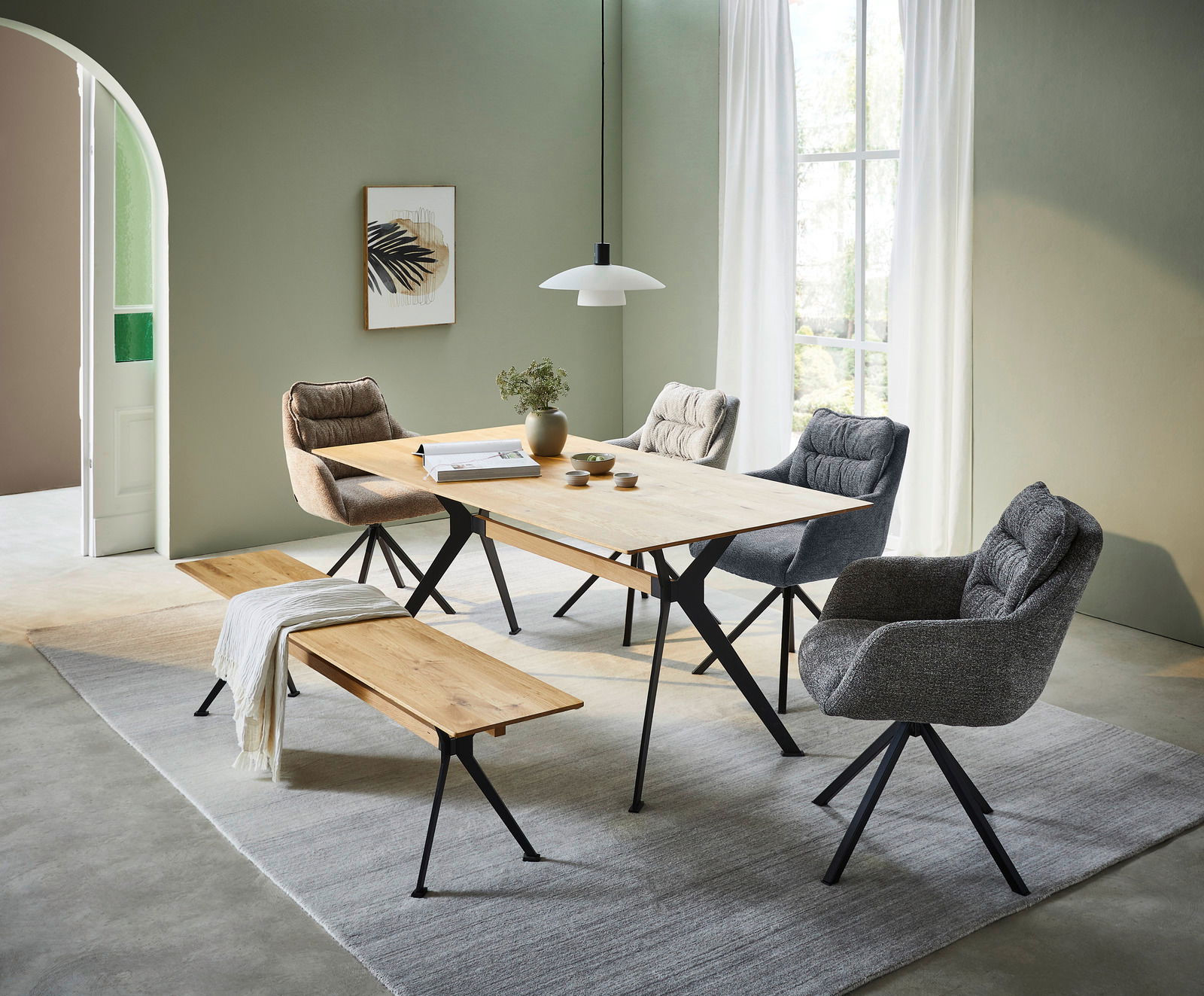 Moderner Esstisch mit Holzplatte und schwarzen Metallbeinen, umgeben von gepolsterten Stühlen und einer Holzbank, in einem hellen Raum mit grünen Wänden und großen Fenstern, seitliche Perspektive.