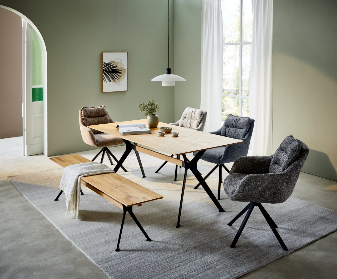 Moderne eettafel met houten blad en zwarte metalen poten, omringd door gestoffeerde stoelen en een houten bank, in een lichte ruimte met groene muren en grote ramen, zijaanzicht.