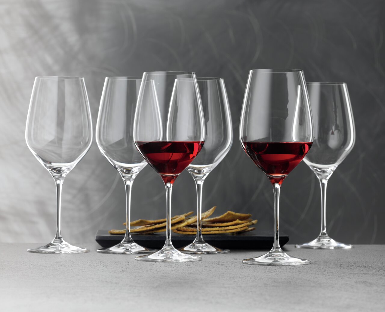 Sechs elegante Bordeauxkelche der Serie Topline, teilweise mit Rotwein gefüllt, auf einer grauen Oberfläche vor einem stilvollen Hintergrund, seitliche Perspektive.