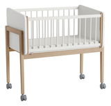 Babybett mit Rollen, Seitenansicht, aus Holz und weißem Material, auf vier Rollen.