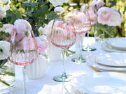 Schräg von vorne fotografierte, elegant geriffelte Weingläser mit zart roséfarbenem Kelch und grünlichem Stiel auf weiß gedecktem Tisch mit Blumenarrangement im Hintergrund.