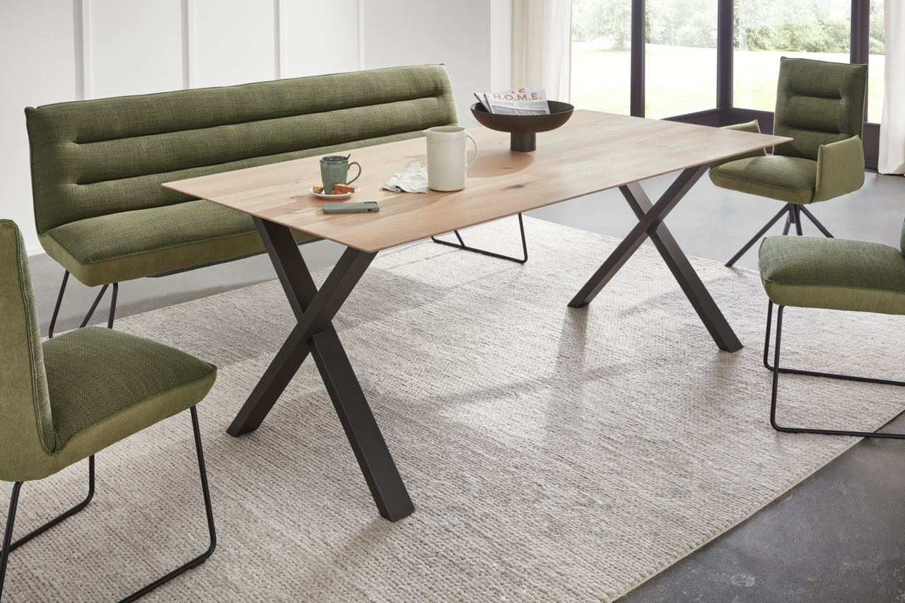 Plantagie Esstisch XAM Moderner Esstisch mit X-förmigen Beinen aus Metall und einer Holzplatte, umgeben von grünen Stühlen und einer Bank, seitliche Perspektive.