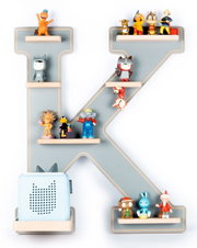 Frontansicht: Großes dekoratives Wandregal in Form des Buchstabens K in Grau mit Holzablagen, bestückt mit kleinen Spielfiguren und einem hellblauen Lautsprecher vor weißem Hintergrund.