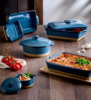 Verschiedene blaue Auflaufformen und Töpfe auf einem Holztisch, gefüllt mit Lasagne und anderen Speisen, aus einer leicht erhöhten Perspektive fotografiert.