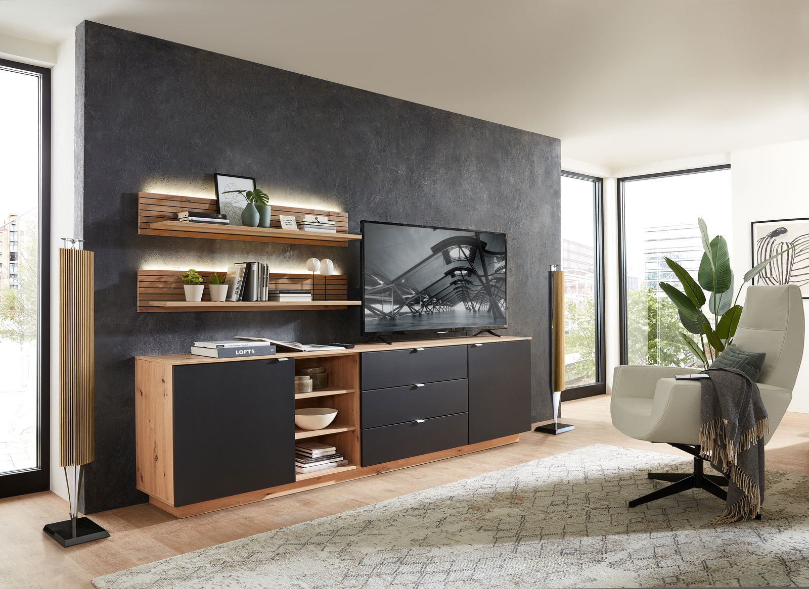 Modernes Wohnzimmer mit Wandboard CORE, das an einer dunklen Wand montiert ist. Das Wandboard ist mit Büchern und Dekorationen bestückt und wird von einer stilvollen Beleuchtung hervorgehoben. Perspektive von der Seite.