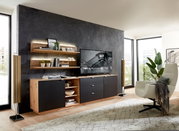 Modernes Wohnzimmer mit Wandboard CORE, das an einer dunklen Wand montiert ist. Das Wandboard ist mit Büchern und Dekorationen bestückt und wird von einer stilvollen Beleuchtung hervorgehoben. Perspektive von der Seite.