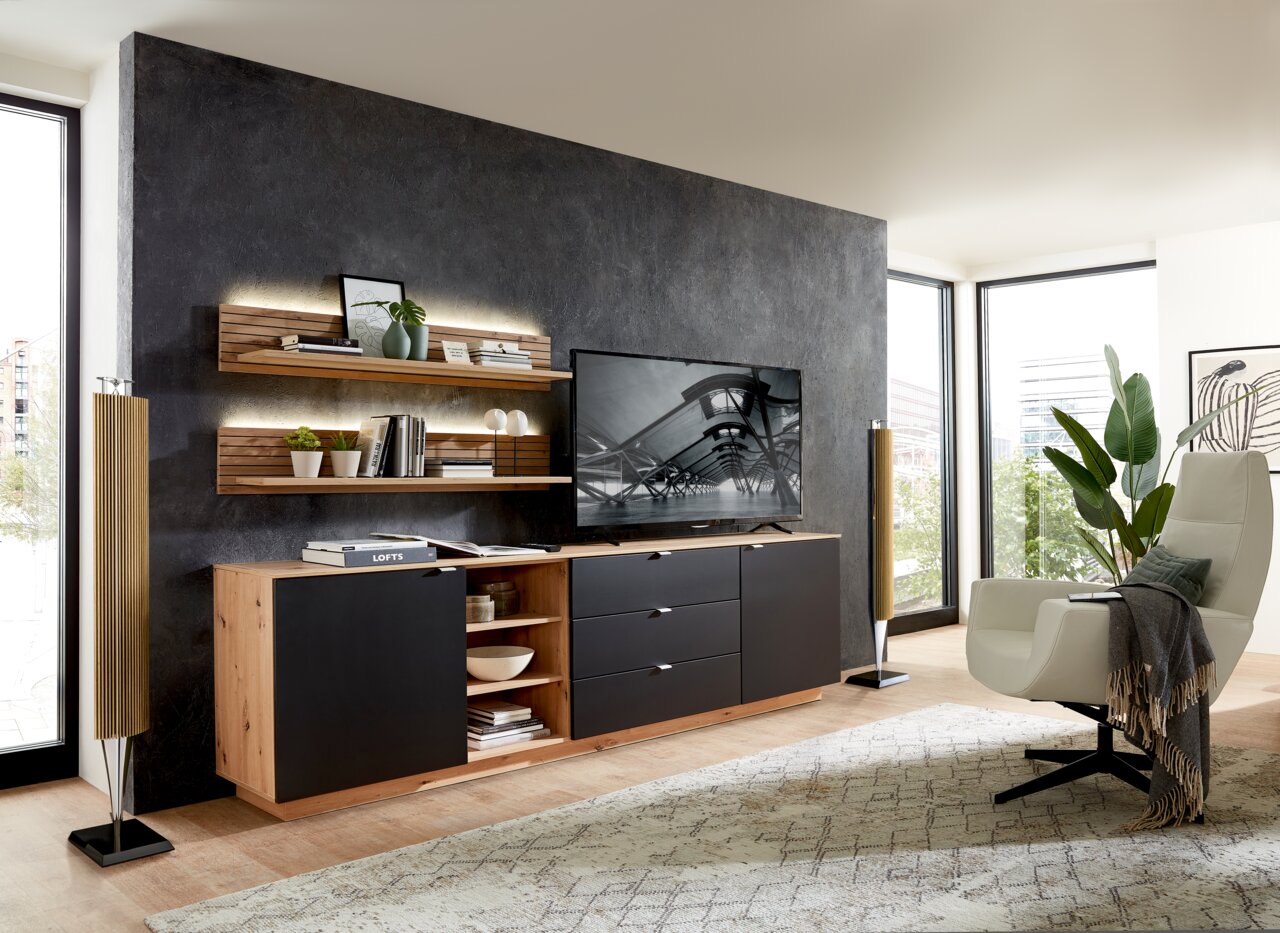 Modernes Wohnzimmer mit Wandboard CORE, das an einer dunklen Wand montiert ist. Das Wandboard ist mit Büchern und Dekorationen bestückt und wird von einer stilvollen Beleuchtung hervorgehoben. Perspektive von der Seite.