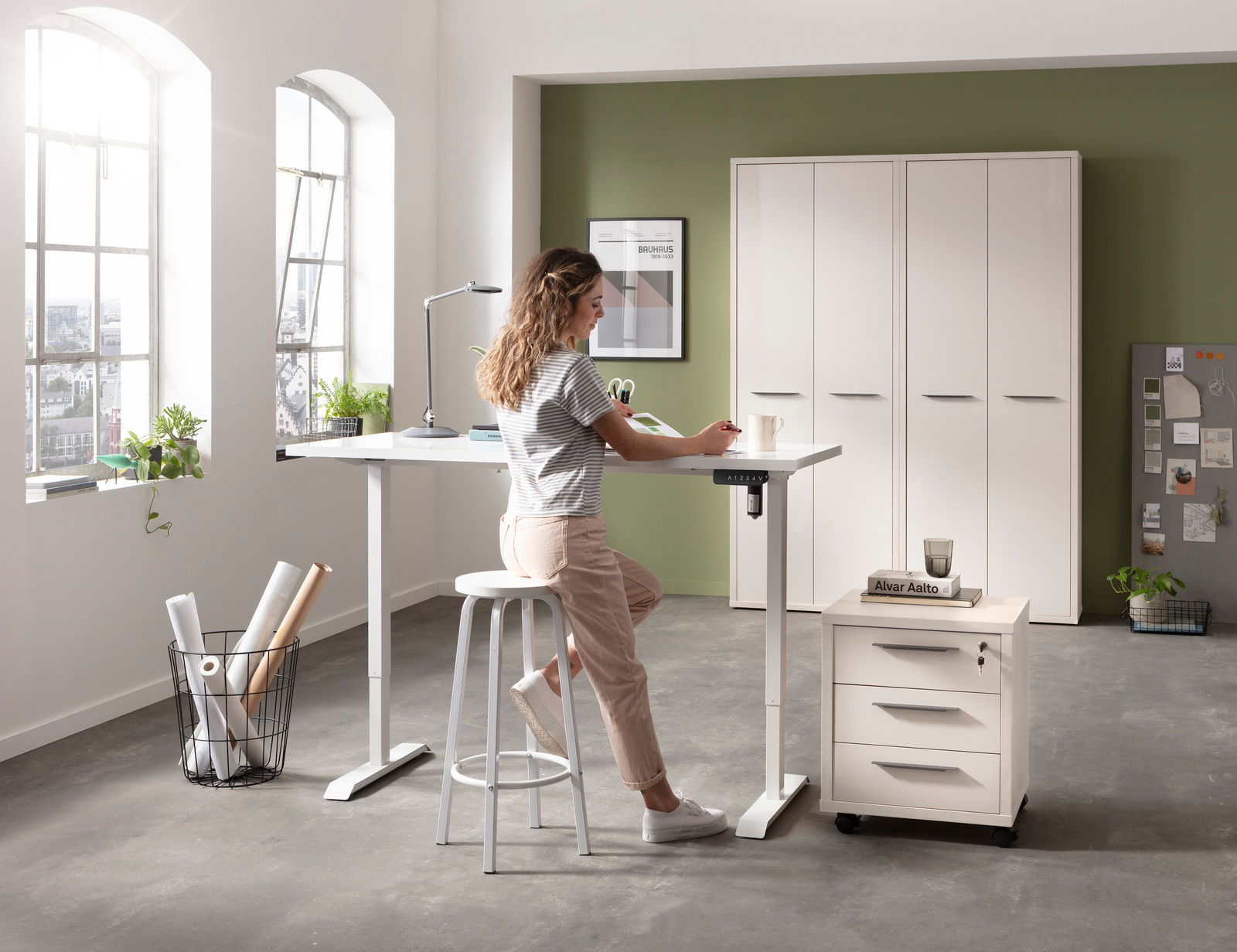 Höhenverstellbarer Schreibtisch mit weißer Tischplatte und elektrischem Fußgestell, seitliche Perspektive, in einem modernen Büro mit großen Fenstern und einer Person, die am Tisch arbeitet.