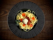 Draufsicht auf einen schwarzen Teller mit Spaghetti, Tomatensauce, geriebenem Käse, halbierten Kirschtomaten und frischen Kräutern auf einem Holztisch