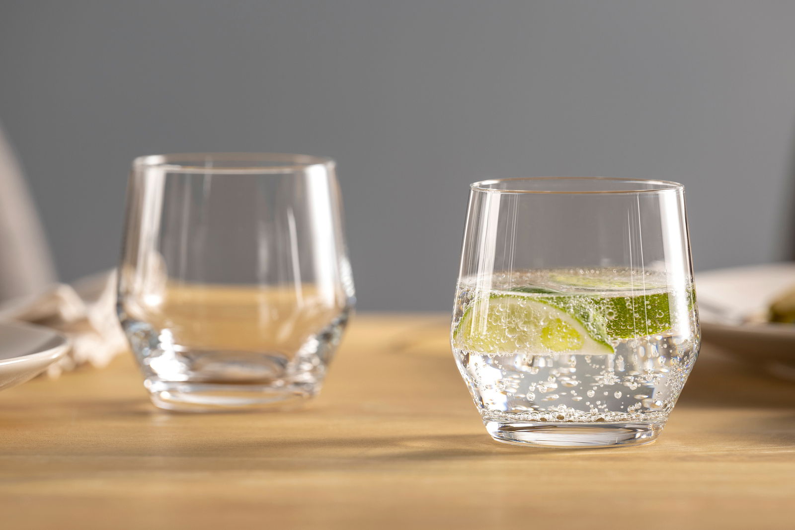 Zwei transparente Becher auf einem Tisch, einer gefüllt mit sprudelndem Wasser und Limettenscheiben, seitliche Perspektive.