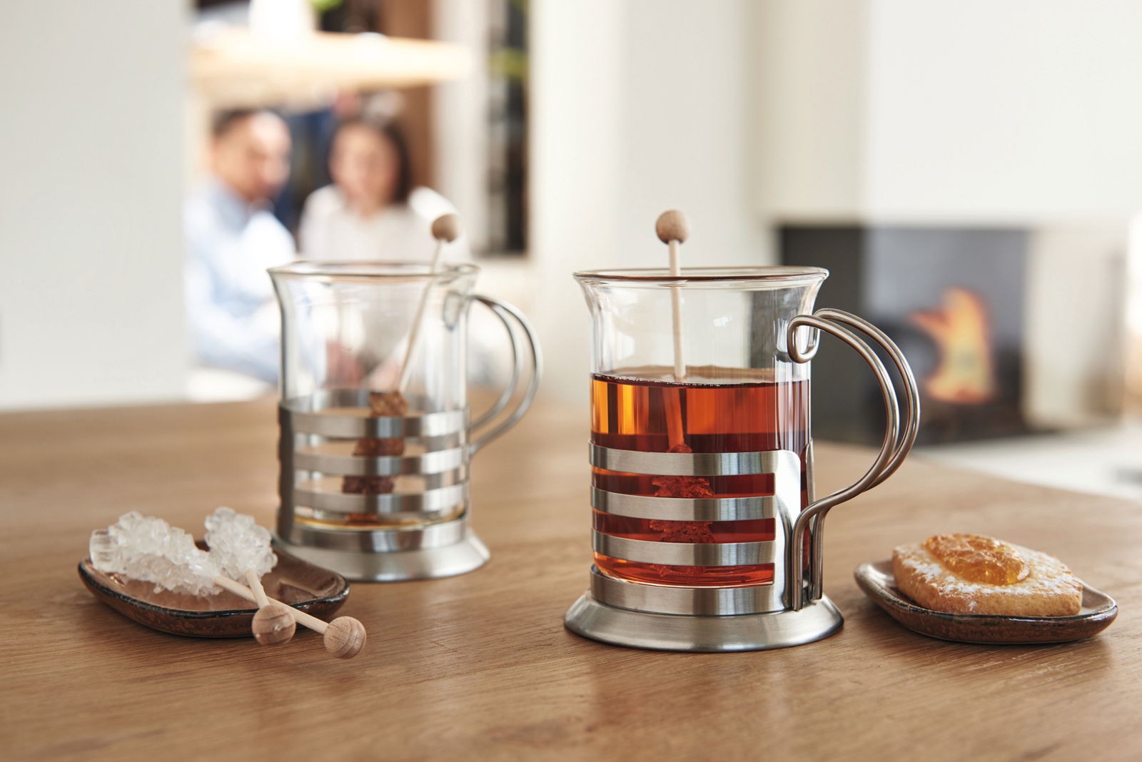 Zwei Teegläser im modernen Design mit Metallhalterung, gefüllt mit Tee, auf einem Holztisch. Im Hintergrund unscharf ein Kamin und zwei Personen. Perspektive von vorne.