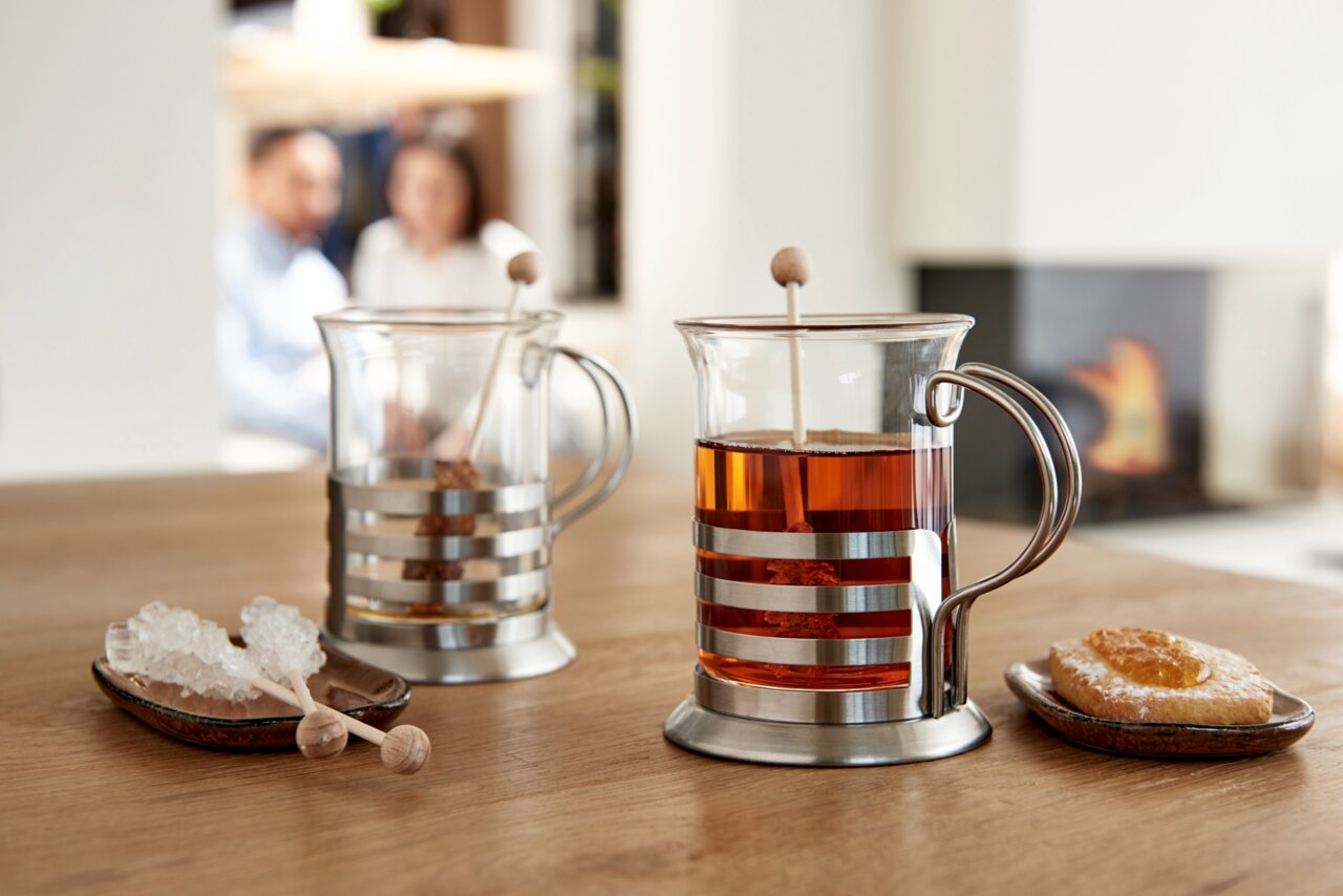 Zwei Teegläser im modernen Design mit Metallhalterung, gefüllt mit Tee, auf einem Holztisch. Im Hintergrund unscharf ein Kamin und zwei Personen. Perspektive von vorne.
