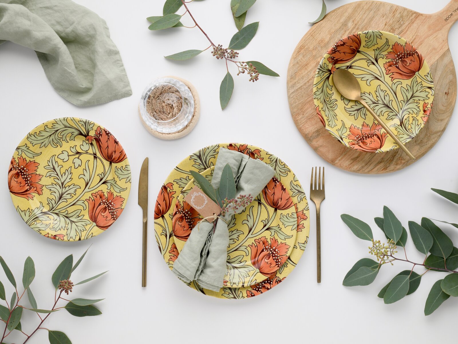 Flaches Top-Down-Foto eines stilvollen Tischgedecks mit gelb-orange floral gemusterten Tellern, goldfarbenem Besteck, hellgrünen Stoffservietten, Holzbrett und Eukalyptuszweigen auf weißem Hintergrund.