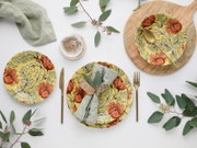 Flaches Top-Down-Foto eines stilvollen Tischgedecks mit gelb-orange floral gemusterten Tellern, goldfarbenem Besteck, hellgrünen Stoffservietten, Holzbrett und Eukalyptuszweigen auf weißem Hintergrund.