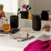 Schwarzer Zahnputzbecher und Seifenspender auf einem Badezimmerwaschtisch, umgeben von dekorativen Elementen und Handtüchern, aus einer frontalen Perspektive betrachtet.