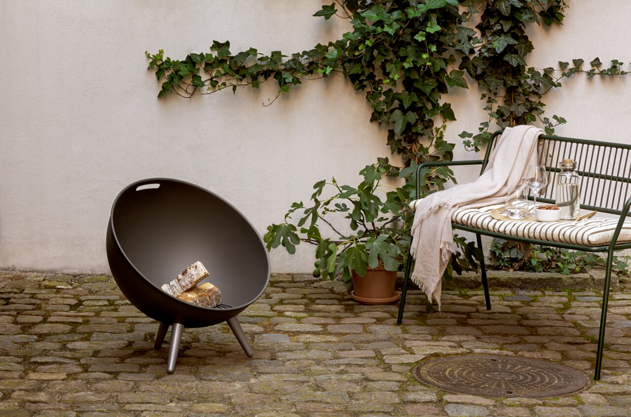 Eva Solo Feuerschale FIRE GLOBE Moderne FireGlobe Feuerschale auf einer Terrasse mit Holzscheiten, umgeben von Pflanzen und einer gemütlichen Sitzgelegenheit, seitliche Perspektive.