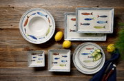 Keramik-Geschirrset mit handgemaltem Fischdekor in Blau, Rot und Gelb, bestehend aus runden Tellern und Schalen sowie rechteckigen Platten, in der Draufsicht auf rustikalem Holztisch arrangiert