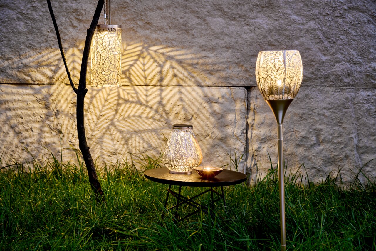 Drei Solarleuchten im Garten bei Dämmerung, eine hängend an einem Ast, eine auf einem Tisch und eine als Stehleuchte, werfen dekorative Lichtmuster auf die Wand.