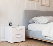 Moderner weißer Nachttisch mit zwei Schubladen, seitliche Perspektive, neben einem grauen Bett in einem stilvoll eingerichteten Schlafzimmer.