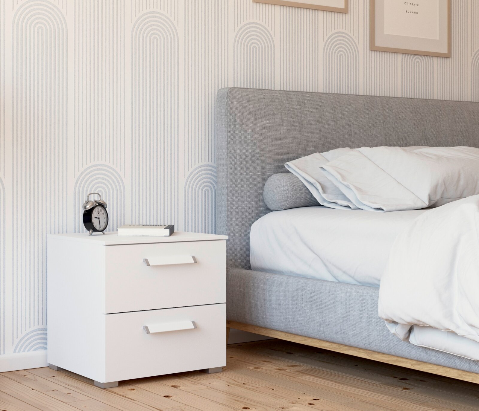 Intro Nachttisch RUDI Moderner weißer Nachttisch mit zwei Schubladen, seitliche Perspektive, neben einem grauen Bett in einem stilvoll eingerichteten Schlafzimmer.