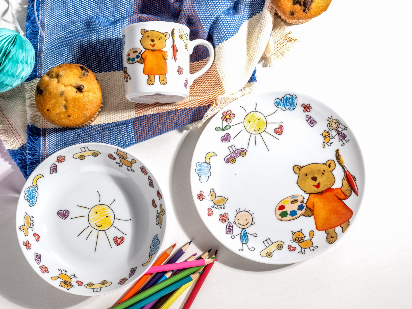 Kinder-Geschirrset aus weißem Porzellan mit Teller, Schale und Tasse, bunt illustriert mit Bär, Sonne und Kinderzeichnungen, fotografiert in Draufsicht von oben auf weißem Hintergrund mit Muffins, bunten Stiften und blauem Tuch