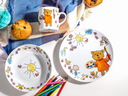 Kinder-Geschirrset aus weißem Porzellan mit Teller, Schale und Tasse, bunt illustriert mit Bär, Sonne und Kinderzeichnungen, fotografiert in Draufsicht von oben auf weißem Hintergrund mit Muffins, bunten Stiften und blauem Tuch