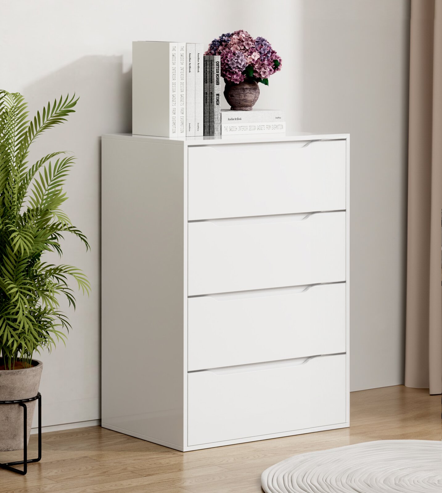 Weiße, moderne Kommode mit vier Schubladen in einem hellen Raum, auf der Oberseite Bücher und eine Vase mit violetten Blumen; Ansicht leicht schräg von vorn.