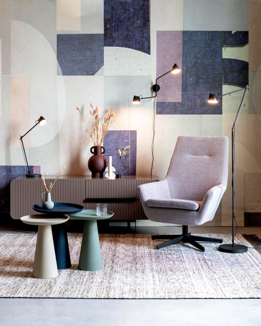 Moderne bijzettafels in verschillende kleuren tegen de achtergrond van een stijlvolle woonkamer met fauteuils en staande lampen, gezien vanuit de zijkant.
