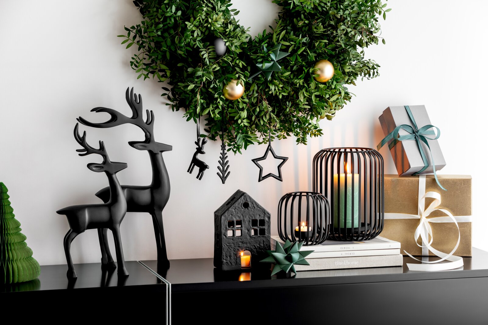 Villeroy & Boch Windlicht Frontansicht einer stilvollen Weihnachtsdekoration mit schwarzen Rentierfiguren, grünem Kranz, leuchtenden Laternen, dekorativem Häuschen und festlich verpackten Geschenken auf einem schwarzen Sideboard.