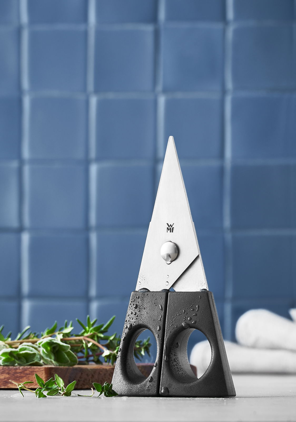 WMF Kräuterschere Kräuterschere 13cm schwarz, stehend auf einem Tisch mit frischen Kräutern im Vordergrund, vor einem blauen gefliesten Hintergrund.